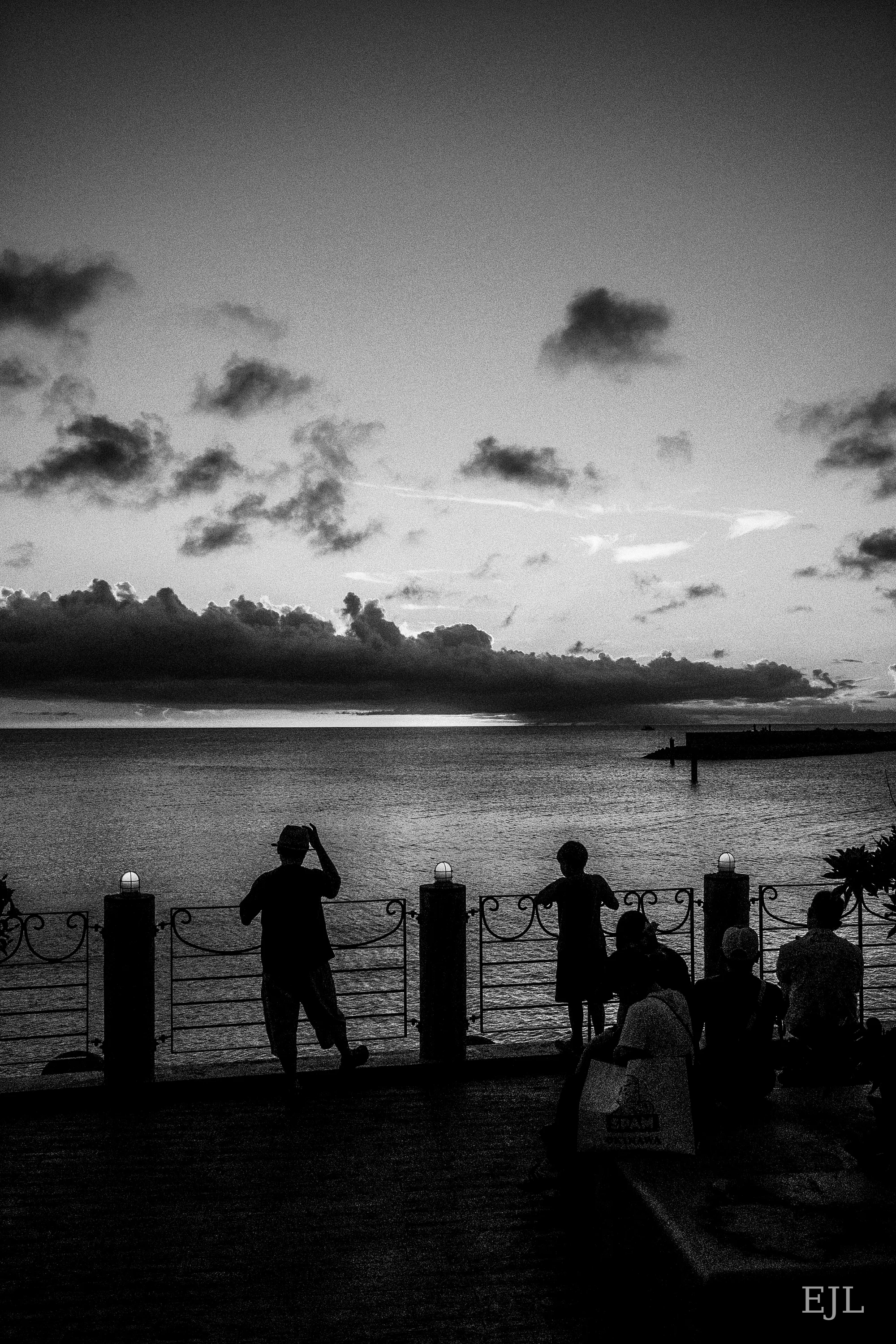 Black-and-white silhouettes of people beside a waterfront railing beneath a cloud-filled evening sky.