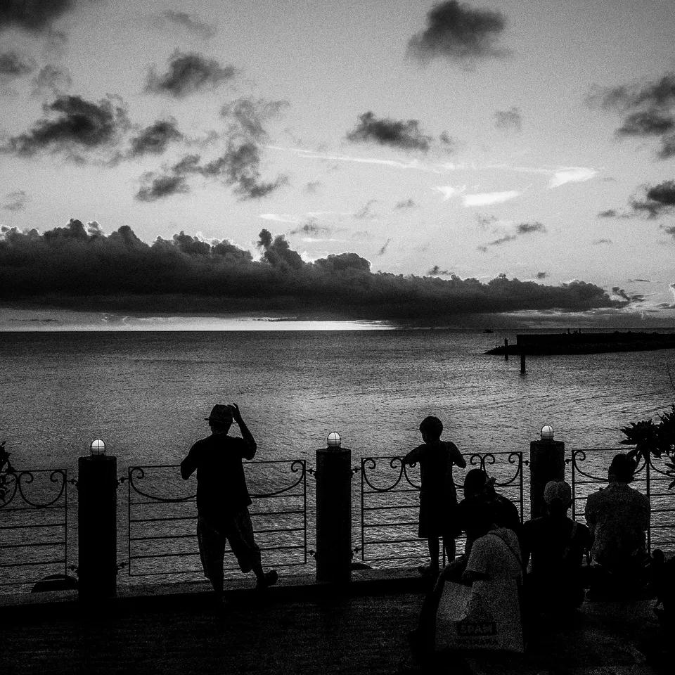 Black-and-white silhouettes of people beside a waterfront railing beneath a cloud-filled evening sky.